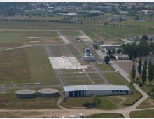 Luftaufnahme eines kleinen Flughafens mit zwei Start- und Landebahnen aus Asphalt und angrenzenden Grünflächen. Im Vordergrund sind runde Tanks und ein Hangar zu sehen, dahinter befinden sich Parkplätze für Kleinflugzeuge sowie Gebäude mit Kontrollturm und Gewerbeeinheiten.