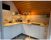 Kompakte Einbauküche mit schräger Holzdecke, weiß gefliesten Wänden und weißen Schränken. Die Küche enthält ein Spülbecken, einen Wasserhahn, eine Kaffeemaschine, einen Wasserkocher, ein Kochfeld sowie offene Regale mit Gewürzbehältern und Büchern.