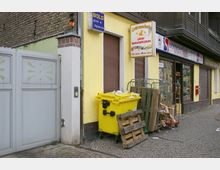 Fassade eines Ladens mit gelber Wand und geschlossenen braunen Rollläden; daneben ein geöffnetes Schaufenster mit Spirituosen. Vor dem Gebäude stehen ein gelber Müllcontainer, Holzpaletten und Metallwagen mit Kartonresten auf einem gepflasterten Gehweg.