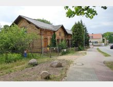 Altes Backsteingebäude mit dekorativen Ziegelmustern an der Fassade, umgeben von Bäumen und Sträuchern, gelegen neben einem Gehweg und einer Straße in einer ländlichen Wohngegend mit weiteren Häusern im Hintergrund.