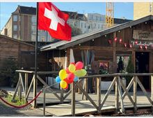 Holzhütte im Chalet-Stil auf einer erhöhten Holzterrasse mit Geländer, davor eine große Schweizer Flagge am Mast und ein Bündel bunter Luftballons. An der Fassade hängen kleine Schweizer Fähnchen und dekorative Skier; im Hintergrund stehen mehrstöckige Stadtgebäude.