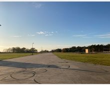 Weitläufiges, flaches Gelände mit gepflastertem Weg in der Mitte und angrenzenden grünen Wiesen, umgeben von Bäumen in der Ferne unter klarem, blauem Himmel. Vereinzelte Laternen und kleinere Holzbauten sind sichtbar.