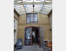 Gelb geklinkertes Gebäude mit einer roten Tür, die von einem Metallgitter umgeben ist, darunter ein rundbogiger Eingang. Darüber befinden sich große, rechteckige Fenster, und das gesamte Areal ist mit einem transparenten Dach aus Glas und Metall überdacht. Im Vordergrund stehen Tische und Stühle auf einem gepflasterten Boden.