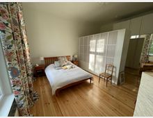 Helles Schlafzimmer mit Holzboden, einem Bett mit Holzrahmen und weißen Bettdecken, farbenfrohen Vorhängen mit Blumenmuster, einem Nachttisch mit Lampe und einer weißen Trennwand im Raum. Im Hintergrund befinden sich ein Schrank mit verspiegelten Türen und ein Stuhl.