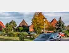 Wohnsiedlung mit mehreren Häusern, die spitze, orange-braune Ziegeldächer haben, umgeben von Bäumen und Sträuchern. Im Vordergrund befinden sich geparkte Autos auf einem Parkplatz, eine Straßenlaterne und gepflegte Grünflächen.