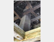 Holzkonstruktion eines Dachstuhls mit massiven, verarbeiteten Balken sowie einem zentralen, gemauerten Pfeiler. Der Blick zeigt die tragenden Strukturen und den Zustand des gealterten Materials unter das Dach.