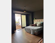 Schlafzimmer mit Holzfußboden, einem Bett mit heller Bettwäsche und mehreren Kissen. Im Hintergrund befindet sich eine offene Glasschiebetür, die zu einer Terrasse mit Holzboden und Gartenmöbeln führt.