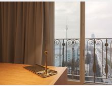 Holztisch mit einer kleinen goldenfarbenen Miniatur des Berliner Fernsehturms im Vordergrund. Im Hintergrund ist ein Balkon mit schmiedeeisernem Geländer und durch die Fenster der Blick auf eine Stadtlandschaft mit dem echten Fernsehturm zu sehen.