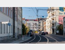 Straßenszene in einem Wohnviertel mit mehrstöckigen Gebäuden links und rechts, Straßenbahn-Schienen in der Straßenmitte und geparkten sowie fahrenden Autos. Im Hintergrund sind historische Gebäude mit Erkern und Giebeln zu sehen, und eine gelbe Straßenbahn fährt in der Ferne.