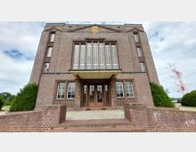 Backsteinbau mit symmetrischer Fassade und hohen Fenstern im Stil der Backsteinexpressionismus-Architektur. Im Vordergrund eine kurze Treppe, die zu einem Eingangsbereich mit Vordach und Doppeltür führt; seitlich Büsche und im Hintergrund ein Teil einer Industriekonstruktion sichtbar.