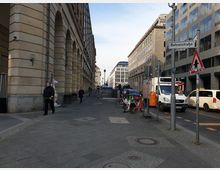 Straßenszene in der Behrensstraße, flankiert von modernen Bürogebäuden und Gebäuden mit historischen Fassaden. Auf dem Gehweg stehen Fahrräder, im Hintergrund sind Autos und ein Transporter zu sehen, neben einem Straßenschild mit einem Warnhinweis.