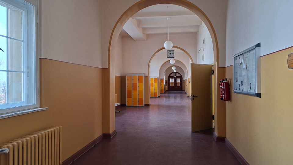 Langer Schulflur mit hohen Rundbögen, glattem Boden und einer Reihe kugelförmiger Deckenlampen, der zu einer Doppeltür am Ende führt. Links sind große Fenster und ein Heizkörper, rechts ein schwarzes Brett und ein Feuerlöscher; entlang des Flurs stehen orange-gelbe Schließfächer.