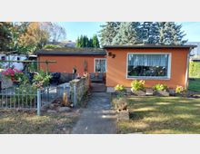 Eingeschossiges, flach gedecktes Haus mit orangefarbener Fassade und großem Frontfenster, davor ein schmaler Betonweg zum Eingang. Links befindet sich ein kleiner, eingezäunter Gartenbereich mit Beeten und Steinmauer, rechts eine Rasenfläche mit Pflanzkästen; im Hintergrund stehen hohe Bäume.