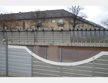 Schallschutzwand mit einer Kombination aus Metall-Paneelen und transparenten Elementen, dahinter ein Wohngebäude aus Backstein mit Satteldach und kahlen Bäumen im Winter.