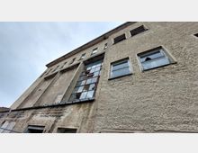 Außenansicht eines alten, verputzten Gebäudes mit großen, vertikal ausgerichteten Fensterflächen und kleineren, rechteckigen Fenstern. Die Fassade wirkt verwittert und zeigt deutliche Spuren von Abnutzung.