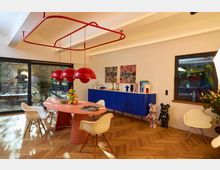 Ein modernes Esszimmer mit hellen Holzböden, einem ovalen rosa Tisch, weißen Stühlen und einer roten Hängelampe. Im Hintergrund stehen eine blaue Kommode, auffällige Dekorationsobjekte, bunte Kunstwerke an der Wand und große Fenster mit Blick auf einen Außenbereich.