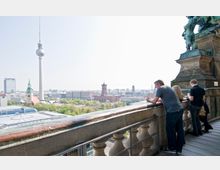 Aussichtsplattform mit Blick auf die Stadt Berlin, einschließlich des Fernsehturms, der Marienkirche und des Roten Rathauses. Im Vordergrund stehen Personen an einem Geländer, während im Hintergrund eine städtische Landschaft mit Bäumen und Gebäuden zu sehen ist.