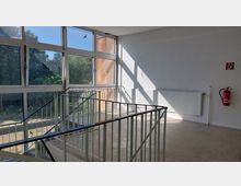 Treppenhaus mit großen Fenstern, die auf eine grüne Außenumgebung blicken. Im Vordergrund ein Metallgeländer, an der weißen Wand ein Heizkörper und ein Feuerlöscher, darüber ein Hinweisschild für den Brandschutz.