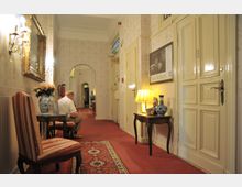 Heller Flur eines Hotels oder historischen Gebäudes mit cremefarbenen Wänden, roten Teppichen und antiken Möbeln. An den Wänden hängen ein goldgerahmter Spiegel und ein Poster, auf einem Beistelltisch stehen eine Tischlampe und dekorative Gegenstände.