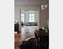 Wohnzimmer in einem Altbau mit hohen Decken und verzierten Stuckelementen, öffnet sich durch eine große Tür zum Essbereich mit Holzboden im Fischgrätmuster; moderne graue Sofas vor großen Fenstern und ein klassischer Holztisch im Vordergrund prägen den Raum.