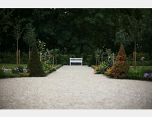 Ein Kiesweg führt durch einen symmetrisch angelegten Garten mit Blumenbeeten und kleinen Bäumen zu einer weißen Holzbank vor einer dichten Baumreihe im Hintergrund.