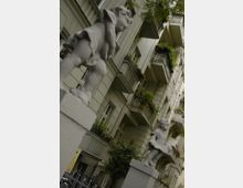 Zwei steinerne Figuren von Kindern stehen auf Säulen im Vordergrund, während im Hintergrund ein mehrstöckiges Wohngebäude mit Balkonen und Grünpflanzen zu sehen ist. Der Gebäudefassade ist hell und durch Fenster sowie verzierte Details geprägt.