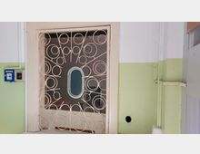 Metallgitter mit dekorativen Kreismustern vor einem Fenster mit ovalem Glaseinsatz in einem Flur mit hellgrün und weiß gestrichenen Wänden; links neben dem Gitter befinden sich eine Hausalarmtaste und ein Rauchabzugshebel.