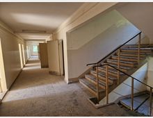 Ein leerer Gebäudeflur mit hellen Wänden und Fliesenboden, auf dessen rechter Seite eine offene Treppe mit Metallgeländer führt. Im Flur sind mehrere geöffnete Türen und ein Fenster am Ende des Gangs zu sehen, durch das Tageslicht hereinfällt.