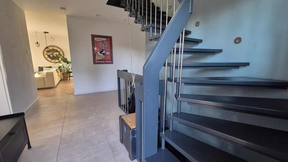 Moderne Inneneinrichtung mit einer offenen Wendeltreppe aus Metall mit dunklen Stufen und silbernen Geländern, auf einem gefliesten Boden. Im Hintergrund ist ein Wohnbereich mit Holzboden, einer hellen Couch, Pflanzen und dekorativen Wandelementen zu sehen.