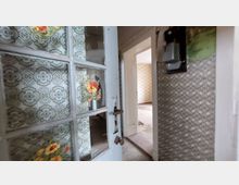 Ein alter, leicht geöffneter Türrahmen mit Glasfenstern, die mit Blumenmotiven verziert sind. Dahinter ein Raum mit verblasster Tapete und abgenutztem Boden, umgeben von weiteren Türen und gemusterten Wänden.