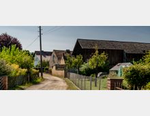 Dorfstraße mit einer unbefestigten Fahrbahn, gesäumt von traditionellen Häusern mit Ziegeldächern und einem großen Holzgebäude. Dazwischen befinden sich Gärten mit Bäumen, Sträuchern, einem Gewächshaus und einfachen Zaunanlagen.