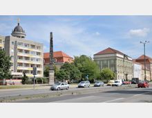 Straßenszene in einer Stadt mit einer Straße im Vordergrund, flankiert von mehreren Fahrzeugen. Im Hintergrund stehen links ein modernes Wohngebäude mit gelben und weißen Balkonen, eine dekorative Säule, sowie rechts historische Gebäude mit grüner und roter Fassade und roten Dächern.