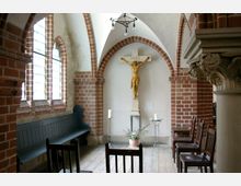 Kapelle mit roten Backsteinwänden und gotischen Spitzbögen, ein Holzkruzifix mit Christusfigur an der Wand, umgeben von Kerzenständern und Sitzbänken aus Holz. Links sorgen hohe, bleiverglaste Fenster für Licht.