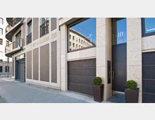 Moderne Gebäudefassade mit hellen Steinflächen und braunen Backsteinsegmenten, großen Fenstern mit Reflexion des gegenüberliegenden Gebäudes und einem großen, dunklen Eingangstor. Zwei symmetrisch platzierte Pflanzkübel mit kugelförmig beschnittenen Büschen flankieren den Eingang.