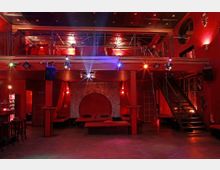 Innenansicht eines Clubs mit rotem Farbschema, hölzernem Boden und Metallverzierungen. Der Raum hat eine erhöhte Galerie mit Geländer, eine Treppe an der rechten Seite und eine Sitzgruppe vor einem dekorativen Wanddesign in der Mitte.