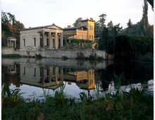 Klassizistisches Gebäude mit Säulen und gelbem Turm in einer parkähnlichen Umgebung, umgeben von Bäumen, mit einem ruhigen Teich im Vordergrund, der das Gebäude und die Vegetation spiegelt.