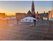 Flachdach mit einzelnen Lüftungsrohren und Dachfenstern aus Kunststoffkuppeln, im Hintergrund ein Kirchturm mit grünlicher Spitze sowie weitere Gebäude bei Sonnenuntergang.