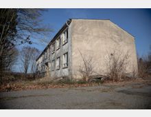 Ein verwittertes, zweistöckiges Wohngebäude mit verblasster Fassade und teilweise beschädigten Fenstern, umgeben von unbewachsenem Boden, kahlen Sträuchern und trockenem Laub. Im Hintergrund sind Bäume und blauer Himmel sichtbar.