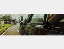 Eine schwarze Limousine mit verchromten Details steht in einer eleganten Auffahrt, umgeben von gepflegten Blumenbeeten und einem schmiedeeisernen Tor. Im Hintergrund ist ein weißes Gebäude im klassischen Stil mit großen Fenstern und Balkonen zu sehen.