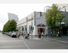 Straßenszene vor einem Gebäude mit dem Schriftzug „Filmtheater“ und einem angrenzenden Imbiss namens „Mister Miller Hot Dog“. Im Vordergrund befindet sich eine Kreuzung mit Fußgängern, geparkten Autos entlang der Straße und Bäumen auf dem Gehweg.