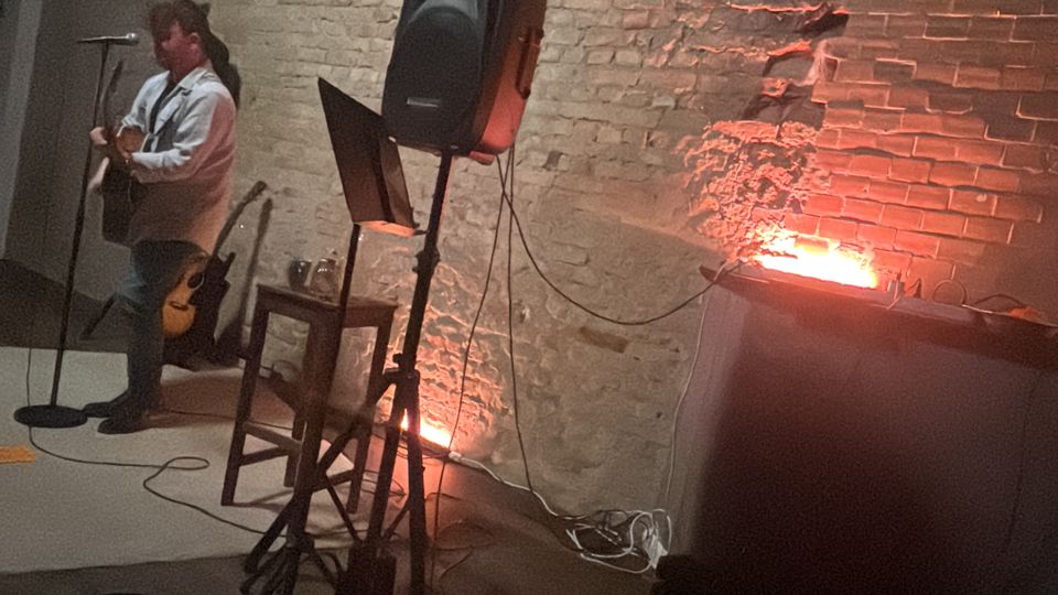 Indoor performance space with an exposed brick wall and open ceiling beams, lit by warm orange uplights. Audio equipment, including a speaker on a stand, microphone stand, mixing console, and cables, sits near a small stage area.