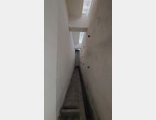 Schmaler, langer Gang mit weißen Wänden und einem Betonboden, der sich nach hinten verjüngt. Oben sind schmale Oberlichter, die diffuses Licht in den Gang werfen.