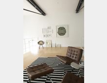 Moderner Wohnraum mit minimalistischer Einrichtung: Zwei braune Lederstühle mit passendem Fußhocker auf einem schwarz-weiß gestreiften Teppich, daneben ein runder Beistelltisch. Im Hintergrund helle Holzböden, eine Wand mit gerahmten Kunstwerken und ein Designerstuhl mit Fellauflage.