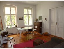 Helles Wohnzimmer mit Holzboden, weißen Wänden und großen Fenstern mit Blick ins Grüne. Im Raum stehen ein Tisch mit Stühlen, ein Sofa, ein Teppich und ein Sideboard mit Deko wie einer Lampe und einem Globus.