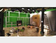 Ein Fernseh- oder Filmstudio im Aufbau mit grünem Hintergrund (Greenscreen), Metallrahmen an der Wand, mehreren Lampen an einer Deckenkonstruktion, Holzvertäfelung an einer Seite und Leitern sowie Kisten auf dem Boden.