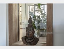 Holzbüste einer asiatisch inspirierten Figur mit geschlossenen Augen auf einem Holzrahmen vor einer Glasfläche; dahinter sind eine große Grünpflanze und ein heller Raum mit Fenstern sichtbar.