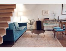 Wohnzimmer mit hellem Holzboden und modernen Möbeln, darunter ein blaues Sofa, ein runder Couchtisch und Regale mit Gläsern. Im Hintergrund befinden sich eine Stehlampe, ein schwarzer Lautsprecher sowie eine Dekovase mit Blumen. Links im Bild ist ein Holztreppenelement sichtbar.