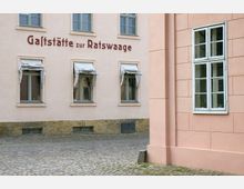 Fassade eines Gebäudes mit der Aufschrift „Gaststätte zur Ratswaage“, hellbeige Wände mit mehreren Fenstern, teils mit weißen Stoffjalousien; gepflasterter Hof im Vordergrund. Rechts im Bild ein angrenzendes rosa Gebäude mit einem Holzfenster.