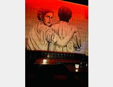 Eine Bar mit gedämpfter roter Beleuchtung, schwarzer Ledersitzbank und dunklen Holztischen. Eine große Wandillustration zeigt zwei Personen in Umarmung, eine davon mit Zigarette, vor einem Hintergrund aus Zeitungsseiten.