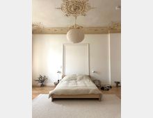 Minimalistisch eingerichtetes Schlafzimmer mit einem niedrigen Holzbett auf einem Teppich. Die Decke ist mit aufwendigen Stuckverzierungen gestaltet, und eine runde Papierlampe hängt zentral darüber.